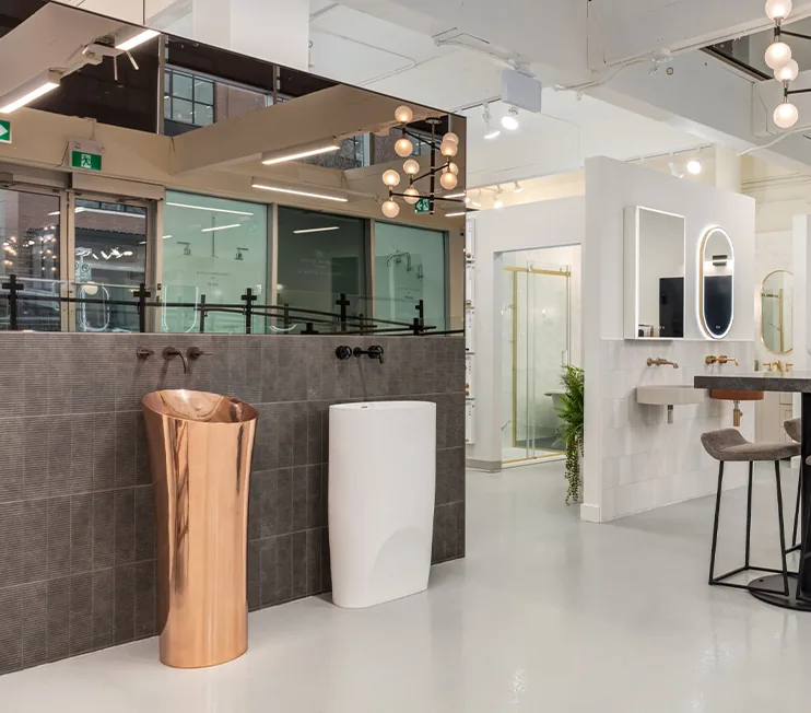 Modern luxury bathroom showroom interior at Baths By Design Calgary featuring freestanding copper and white pedestal sinks, contemporary wall-mounted faucets, illuminated mirrors, and glass shower displays.